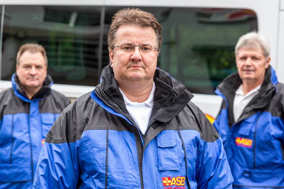 Uwe Köhler,Fachdienstleiter Fahrdienst,steht zwischen zwei Mitarbeitern seines Teams. Alle drei Männer tragen blaue Jacken mit dem ASB-Logo