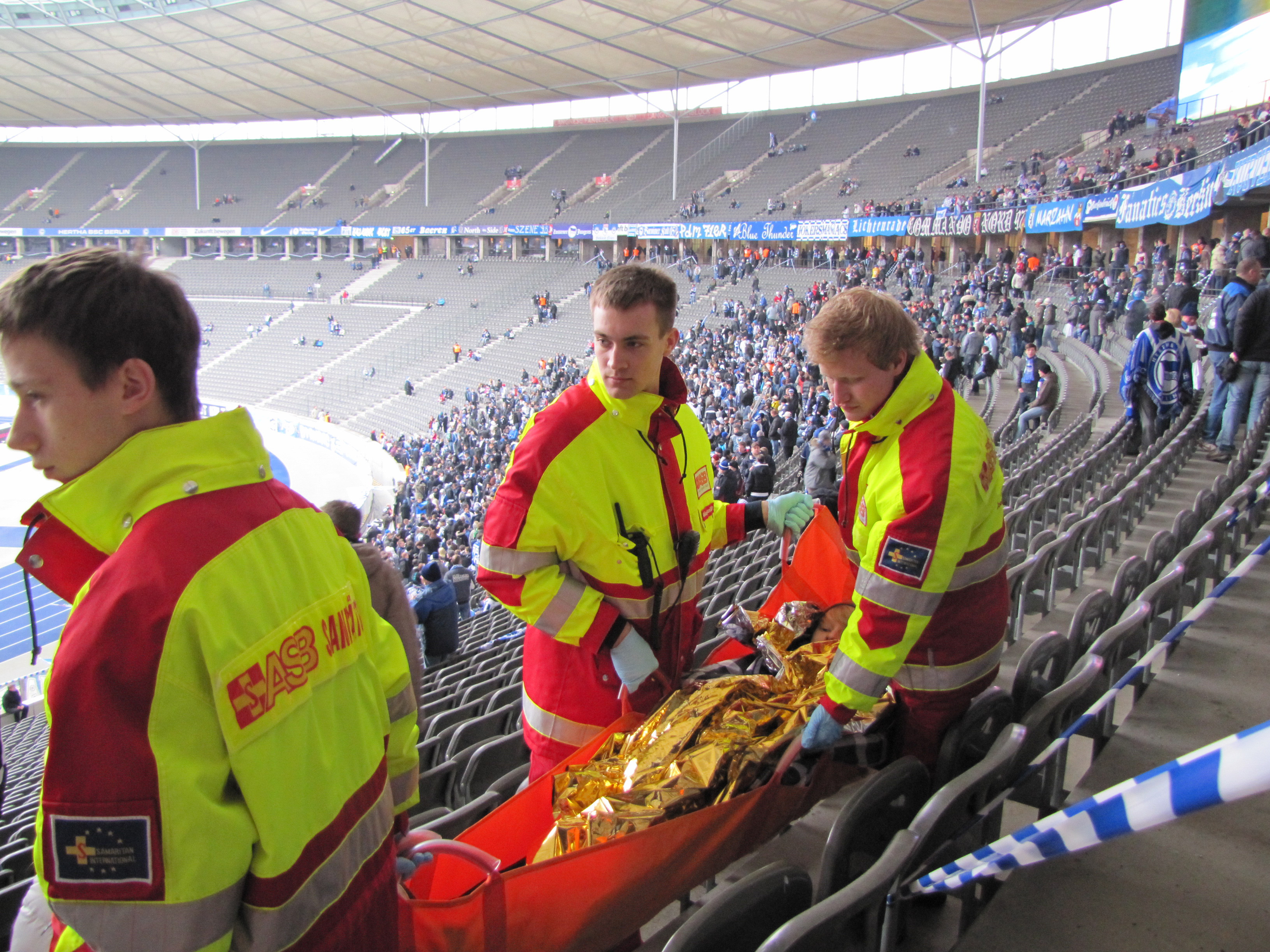 Drei ASB-Sanitäter laufen mit einer Trage durch die Ränge eines Fußballstadions. Im Hintergrund sind viele Fußballfans zu sehen